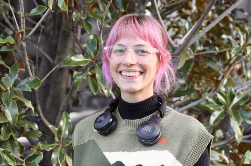 A woman with pink hair and clear-framed glasses smiles directly into the camera. 