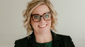 A professional headshot photo of Annie Kubes. She has short, blond, wavy hair, is wearing brown square glasses, and is smiling in the photo. She is also wearing a blazer over a green blouse. 