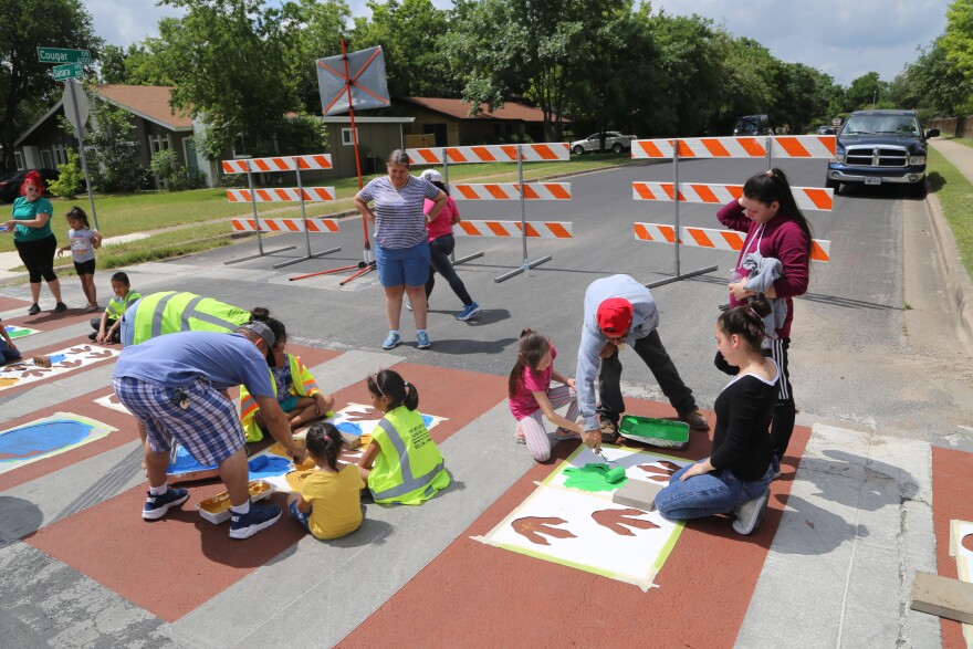 Adults helping children to paint stencils on a crosswalk. Safety barriers are set up to prevent cars from driving through.