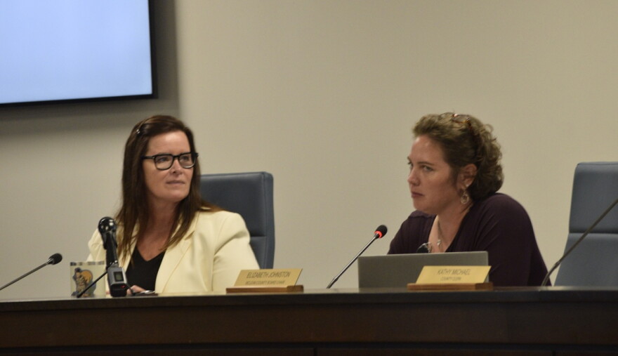 Two women sit at a desk with microphones. 