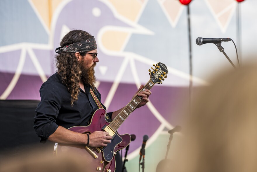 A guitar player on stage with a maroon guitar. He has long curly brown hair held back by a black bandana headband.