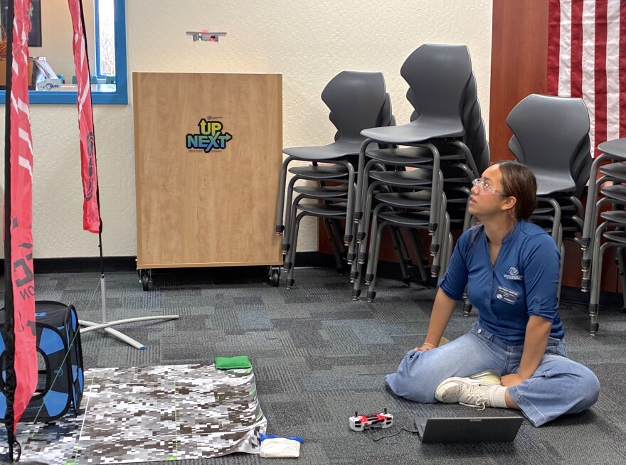 A young woman, sitting on the floor next to an open laptop and drone controller, watches a small drone float beneath a pink arch.