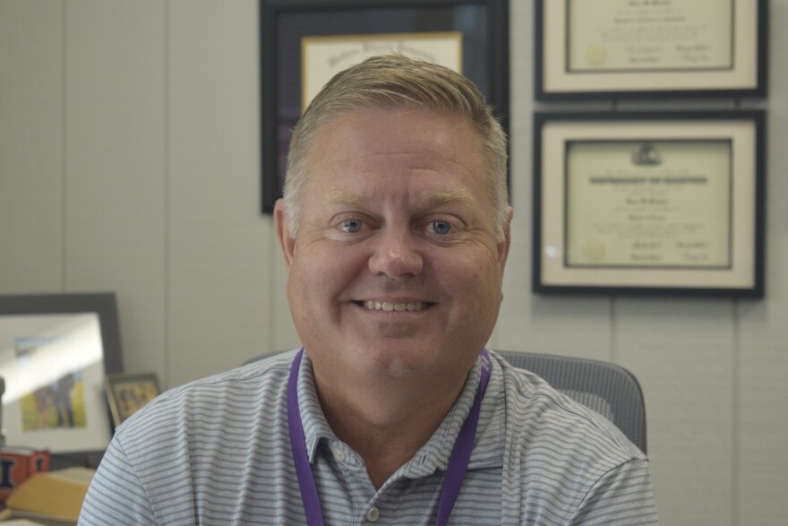 A middle-aged man with short blond hair and a striped polo shirt sits smiling in an office. Framed certificates and photos are visible on the wall behind him.