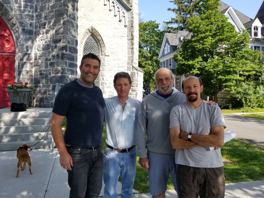 Four men stand in front of a stone church with a dog in the background