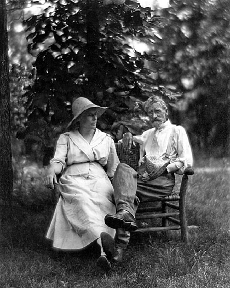 A black and white photograph of artist T. C. Steele and his wife Selma Neubacher Steele sitting outdoors on a wooden bench or chair. Selma, wearing a light-colored dress and sun hat, sits on the left, looking toward Steele. Steele sits on the right, wearing a light-colored shirt, suspenders, and boots, with one leg crossed over the other and his foot resting on the bench. They are seated in a grassy area with dense foliage behind them. 