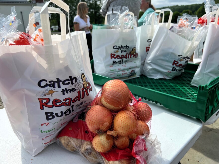 Bags of onions and potatoes sit stacked on a white folding table, in front of another bag filled with bread.