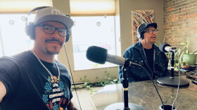 Two men in a radio studio sitting behind microphones. One is holding his arm out to take the selfie.