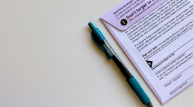 A mail-in ballot envelope with a pen sitting beside it. Some of the info on the ballot is visible, such as a reminder to sign the ballot and voting laws.