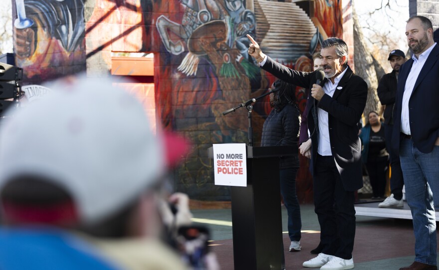 A man stands at a podium holding a microphone and gesturing to a crowd. Other people stand behind him.