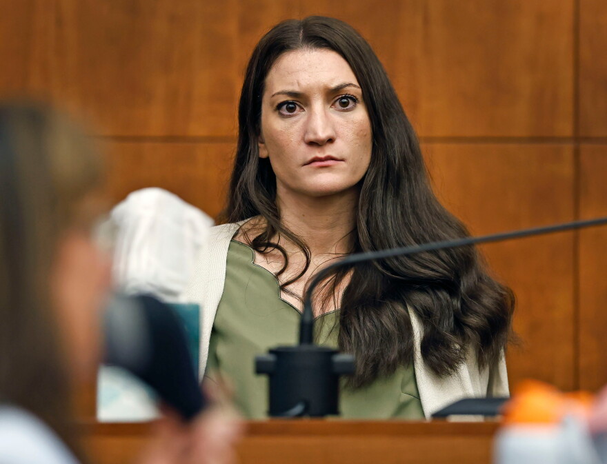 A woman with long brown hair sits on a witness stand in a courtroom, looking serious. The background is wood-paneled, and a microphone is in front of her.