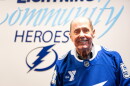 Older bald man wearing blue hockey jersey and smiling into the camera with a logo of a lightning bolt behind his right shoulder