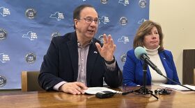 State Sen. Joseph Griffo (R-53), and Assemblywoman Marianne Buttenschon (D-119) sit at a news conference table talking to the press. A blue banner is behind them with the New York State logo and seal behind them.
