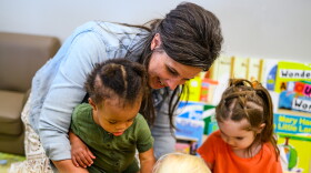 A woman smiles as she interacts with three young children in a classroom, surrounded by colorful books and educational materials.