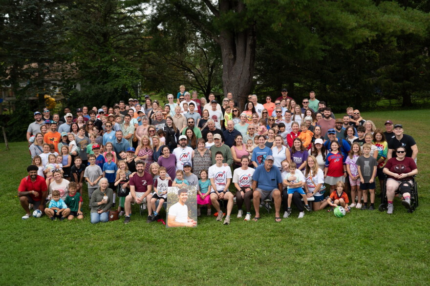 This is a group of people posing in a park in front of a tree. They have a poster with a picture of Nick Henry, who passed away after a battle with cancer. 