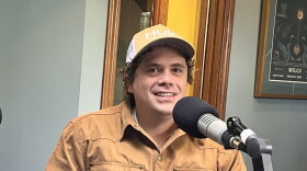 A man in a tan jacket and yellow baseball cap sits behind a microphone in the KAXE studio.
