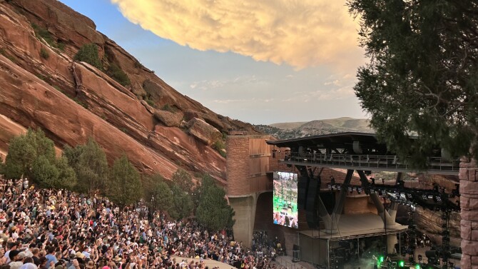 Music fans enjoy a sunset concert at Red Rocks Amphitheater. On the left side is large, tilted rock formation that is a rust-red color with green vegetation. Thousands of fans are seated on a downward slope facing the stage. The band performs on the stage in the lower right corner. There is a big green tree, the sky is blue and sun is setting behind a big cloud.
