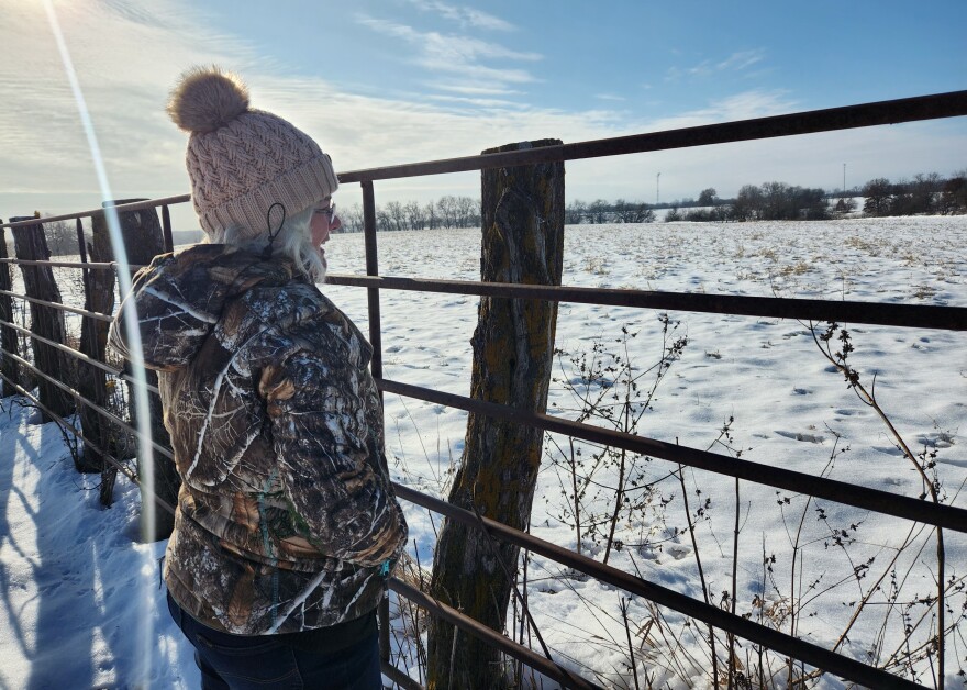 A woman in a camouflage jacket and hat stands near a white snowy field.