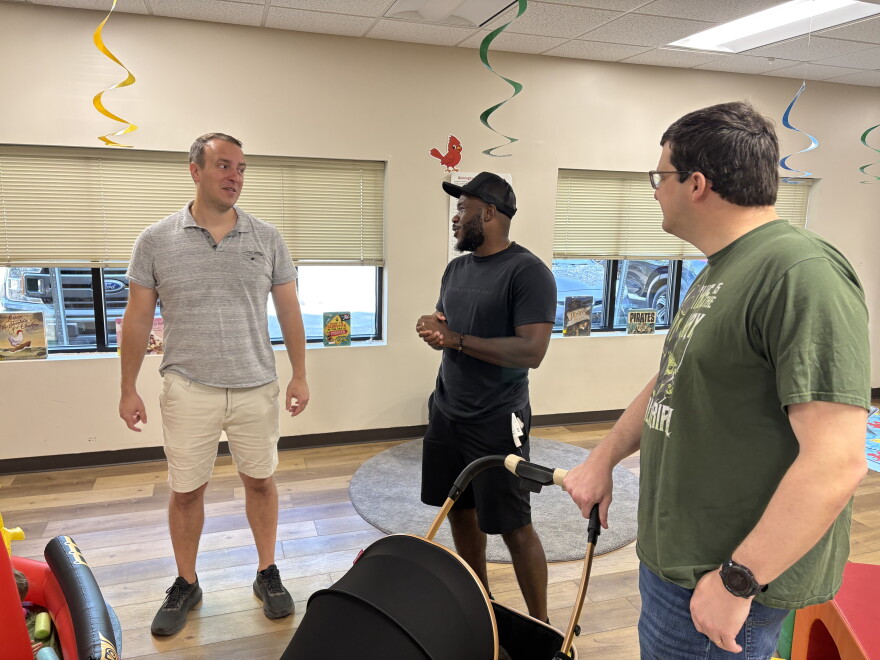 Three men stand and talk. One hold the handle of a stroller.