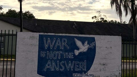A white slab brick wall leans against a black fence; a white building and palm trees are visible in the background. The sign reads: "WAR IS NOT THE ANSWER" in dark blue, with a white dove carrying an olive branch at the edge.