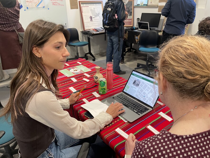 A woman listens to another woman as they sit in front of a computer. The computer is on a table with a red table cloth and candles on it.