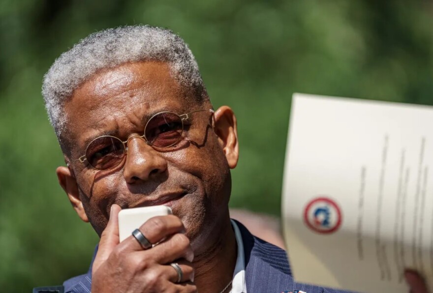 Allen West speaks into a handheld microphone while holding a piece of paper with the GOP logo on it.