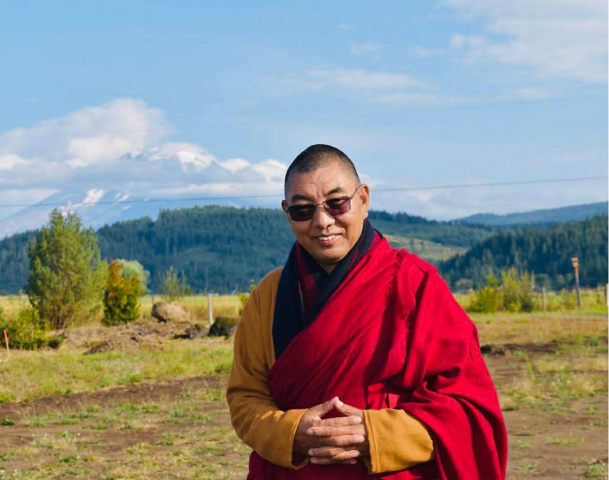 A buddhist monk smiles at the camera with his fingers interlocked with mountains in the background