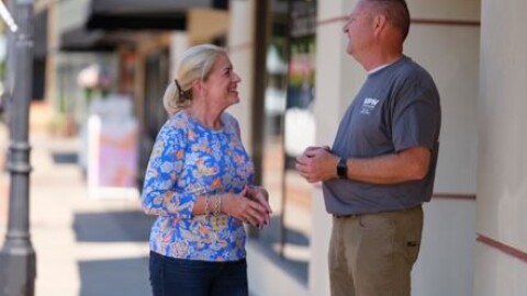 A woman and man stand on a sidewalk smiling towards each other. 
