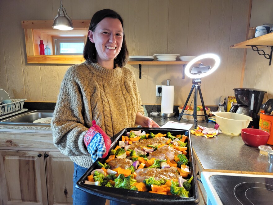 Mackenzie Sachs holds a baking sheet with chicken and vegetables in a kitchen.