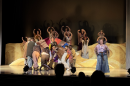 A man wears a colorful long coat that other actors hold as they circle him. Dancers and a narrator stand on stage singing.