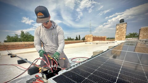 A man in a grey shirt on a roof next to a black solar panel and red electrical chords.