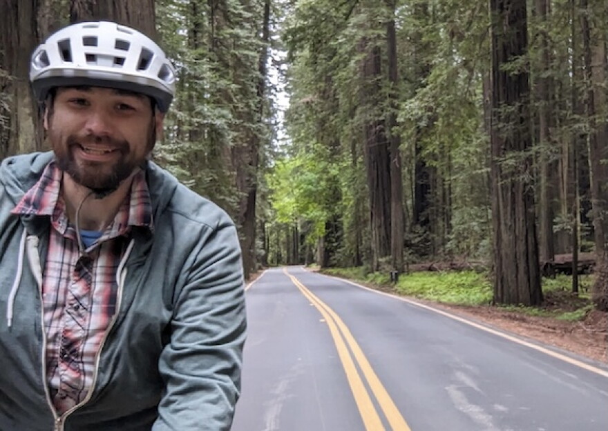 Andrew Wolf wears a bicycle helmet while standing beside a two-lane road surrounded by tall trees.