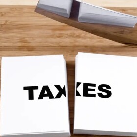 illustration shows an ax next to a stack of cards cut in half with the word TAXES written on the top card. Illustrates elimination or trimming of taxes 