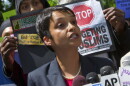 A young woman with short dark hair and dark complexion in a gray blazer and pearl like necklace speaking behind microphones with people behind her holding up signs about targeting muslims. 