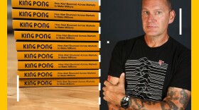 A stack of books and the author Raiford Guins stand next to each other, the screen has the overlay of a game of Pong on it.