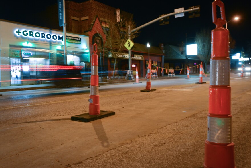 Nighttime scene showing street pavement in the foreground with tall, orange pylons placed on the street to delineate lanes for traffic. The red tail lights of a car are blurred as it passes in front of businesses in the background.