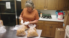 A woman with gray medium length hair in a short sleeve dark orange t-shirt twists a tie onto a bag of pretzels. Three other bags sit on the metal table in front of her. Behind her is a cluttered kitchen countertop at the back of the small room, which has cabinets lining the wall above and below it. The counter has a few different items on it, including three dark green thermoses and a red and white bag of all-purpose flower. To the right of the image can be seen a black fridge behind the woman.  