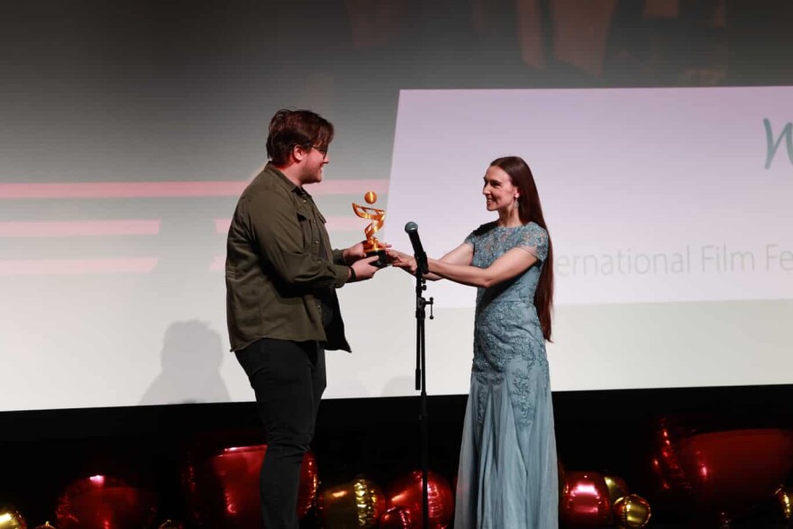Two people stand on a stage behind a microphone. One person hands another an award.