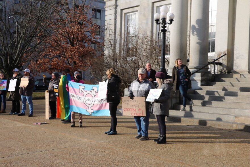 A photo of several people in front of the State House, one holding a trans rights flag. 