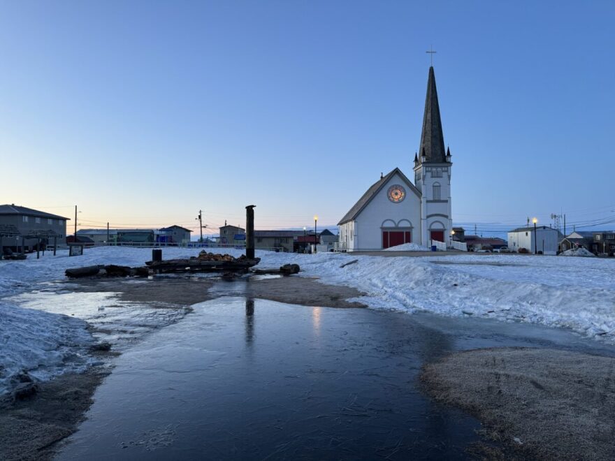 The Iditarod Trail’s famous Burled Arch rests in pieces scattered across the ground with Old St. Joe’s Church in the background. One of the pillars of the arch lies on the ground.