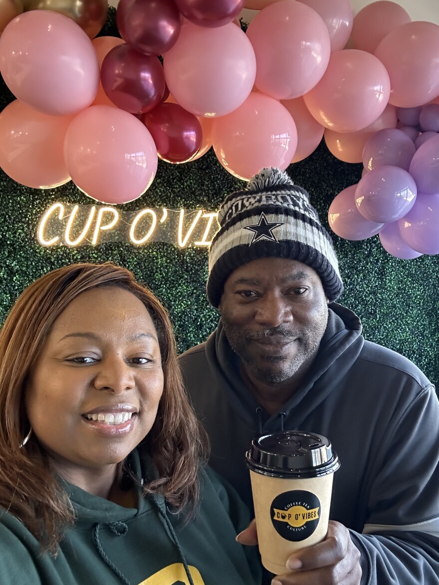 Charletra Sharp, in dark green Baylor University colors, poses with a drink from her coffee shop, Cup O' Vibes with a man on her right in a Dallas Cowboys beanie. Behind them hangs a green floral backdrop and a neon sign bearing her Arlington business name.