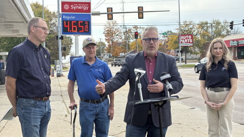 A man in a blazer speaks at a podium with two microphones, flanked by a man in a blue polo, an older man with a cane, and a woman in a black polo, with a gas station price sign in the background.