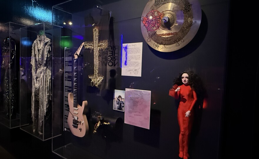 A glass museum case displays a pink guitar, a black tank top with a cross decal made of bobby pins, a photograph of a woman, a black flamethrower megaphone, a doll with long black hair wearing a red bodysuit, two pages of songwriting notes and a cymbal covered in signatures.