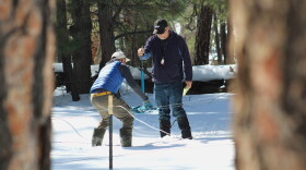 Two men stand in a snowy forest, holding a system of tubes and string and reading measurements from the tool