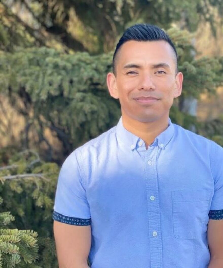 a hispanic man standing for the camera in front of a tree with a blue button up shirt