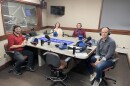 Four people wearing headphones sit at a table in a radio talk studio: a man front left has short dark hair, a dark beard, and is wearing glasses, a red shirt with a blue collar, black pants, and black shoes; a man front right has short dark hair and is wearing a grey button-down shirt, jeans, and sneakers; a woman back left has strawberry blonde hair and is wearing a black and white shirt under a cream-colored cardigan; a man back right has his long grey hair in a ponytail, a grey goatee, and is wearing a maroon button-down shirt.