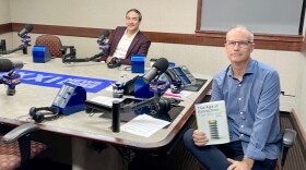 Two smiling men sitting at a table in a radio talk studio: the man back left has short dark hair and a dark mustache and is wearing a white button-down shirt under a burgundy suit; a man front right has very short dark hair and is wearing a blue button-down shirt, jeans, blue socks, and white and red sneakers. He is holding a book titled "The Age of Extraction." 