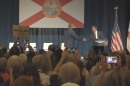 Florida Gov. Ron DeSantis shakes hands with U.S. Rep. Byron Donalds onstage in front of a large Florida flag as audience members watch and take photos.
