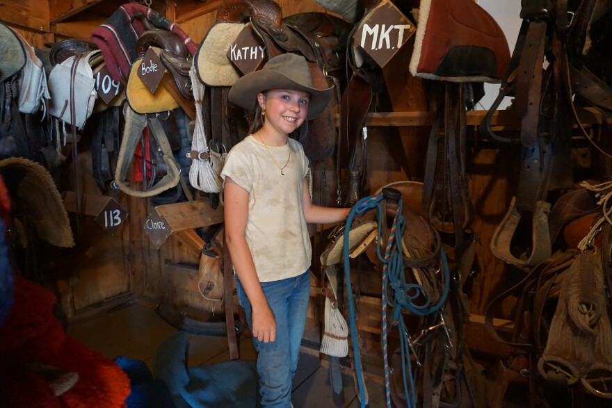 A young girl smiles, wearing a cowgirl hat and holding a saddle. She stands in front of a wall of other horse saddles.