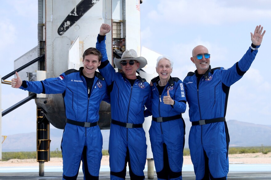 Four people in blue spacesuits pose with smiles, outstretched arms and thumbs up in front of a desert landscape.