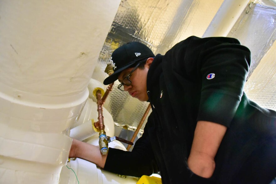 A high schooler in a black hooded sweatshirt and hat works on large, white pipes.